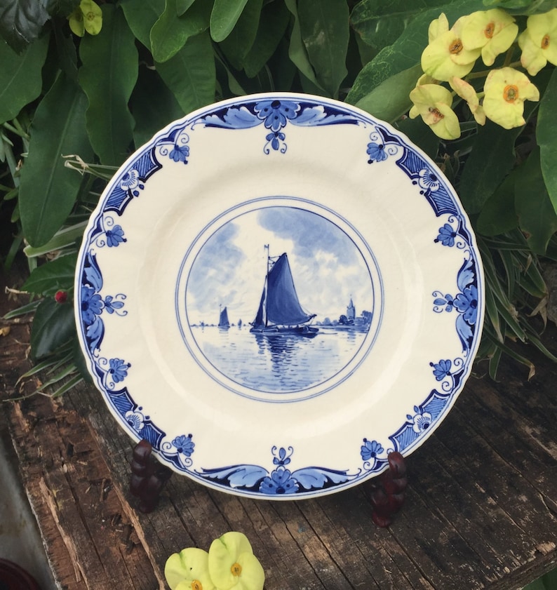 May include: A white ceramic plate with a blue and white design featuring a sailboat on a body of water. The plate has a scalloped edge and a blue and white floral border.