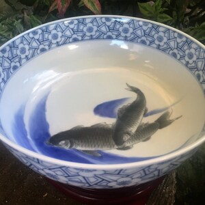 K&ouml;nnte beinhalten: Eine wei&szlig;e Keramikschale mit blauem Blumenmuster am Rand und einem zentralen Design von drei grauen Fischen, die in blauen Wellen schwimmen. Die Schale steht auf einem dunkelroten Holzsockel.