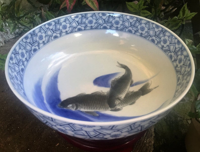 K&ouml;nnte beinhalten: Eine wei&szlig;e Keramikschale mit blauem Blumenmuster am Rand und einem zentralen Design von drei grauen Fischen, die in blauen Wellen schwimmen. Die Schale steht auf einem dunkelroten Holzsockel.