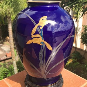 May include: A cobalt blue ceramic vase with a gold rim and a black base. The vase features a gold floral design with silver accents. The vase is a decorative item, suitable for displaying flowers or as a standalone art piece.