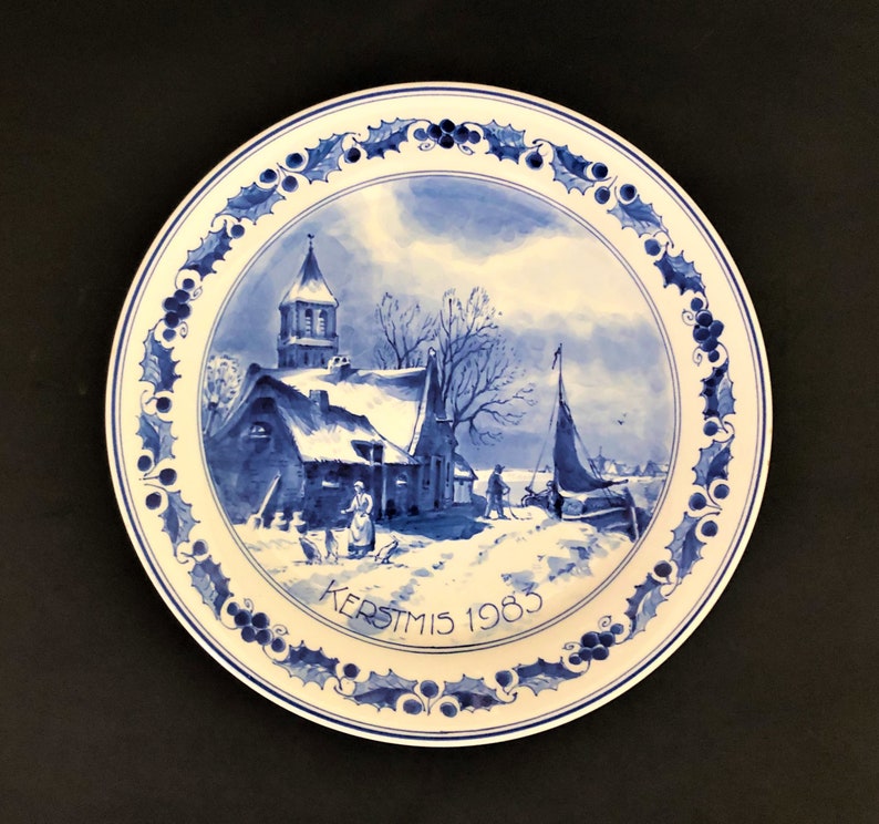 May include: A round, white ceramic plate with a blue and white winter scene. The plate features a church, figures, and a sailboat. The rim has a blue holly berry design. The text "KERSTMIS 1983" is written on the plate.