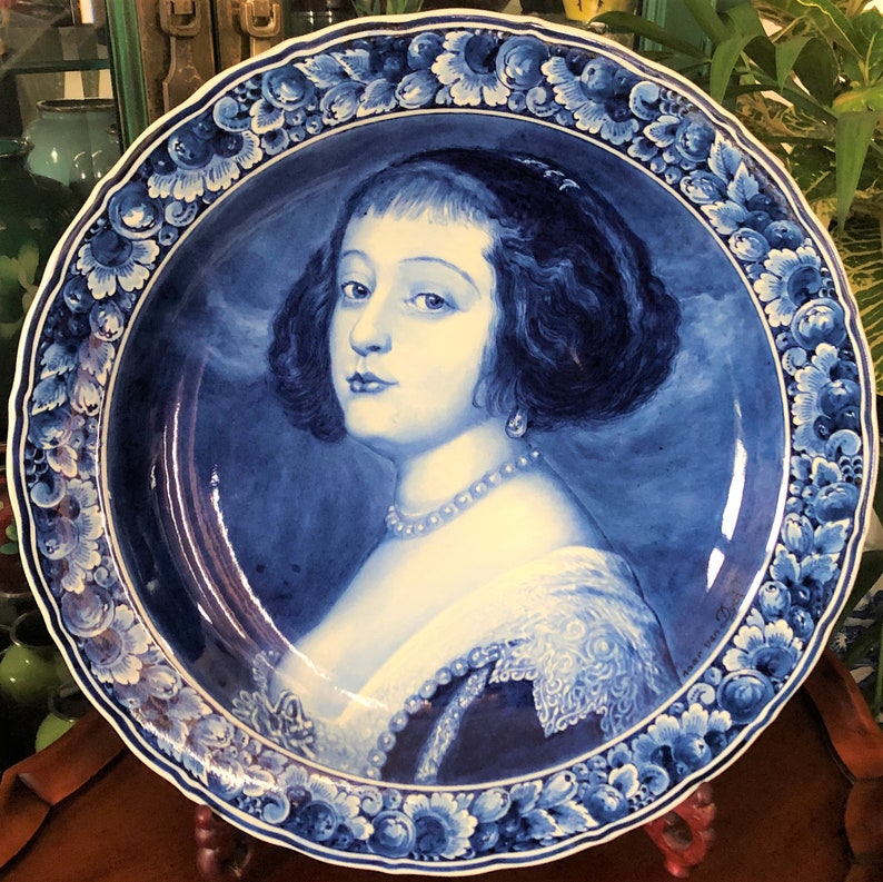 Pu&ograve; includere: Un piatto in maiolica di Delft blu e bianco con un ritratto dipinto di una donna con i capelli scuri e una collana di perle. Il piatto &egrave; decorato con un bordo floreale.