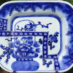 Op de afbeelding: Een vierkant blauw en wit porseleinen bord met een bloemenmotief. Het bord heeft een blauw en wit bloemenmotief met een vaas en een rand van dennennaalden.