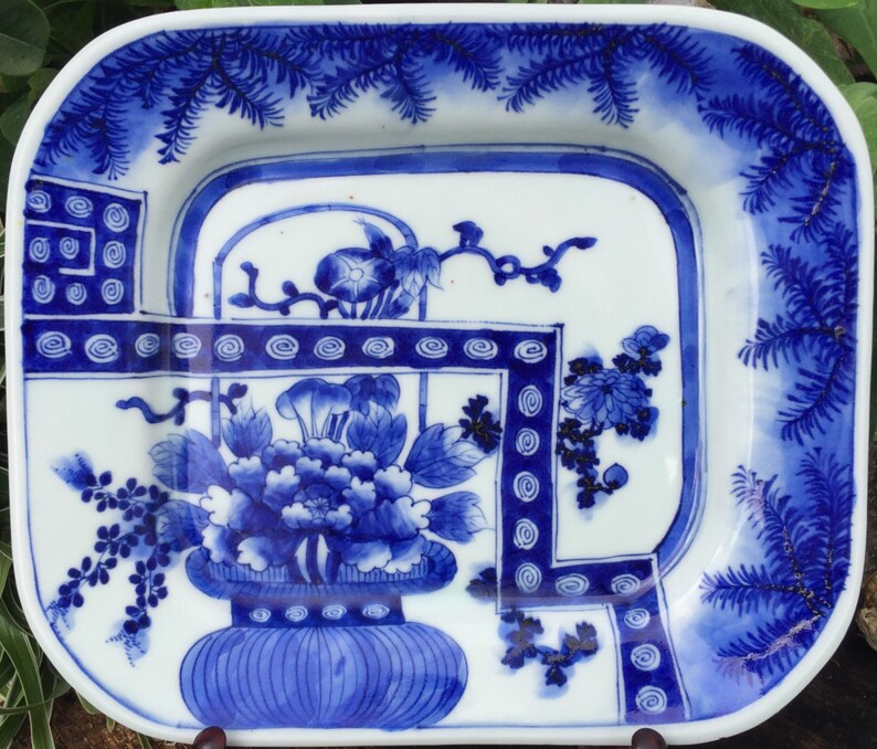 Op de afbeelding: Een vierkant blauw en wit porseleinen bord met een bloemenmotief. Het bord heeft een blauw en wit bloemenmotief met een vaas en een rand van dennennaalden.