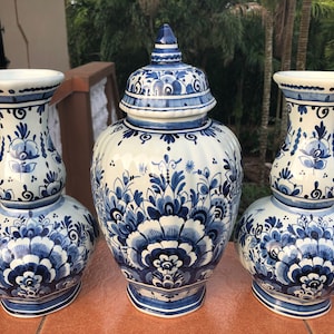 May include: A set of three ceramic vases with a blue and white floral design. The set includes two vases with a flared top and a lidded jar. The vases have a bulbous base and are decorated with intricate patterns.