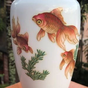 Pu&ograve; includere: Vaso in porcellana bianca con bordo argentato e tre pesci dorati dipinti sul davanti. I pesci nuotano in un disegno floreale verde e bianco.