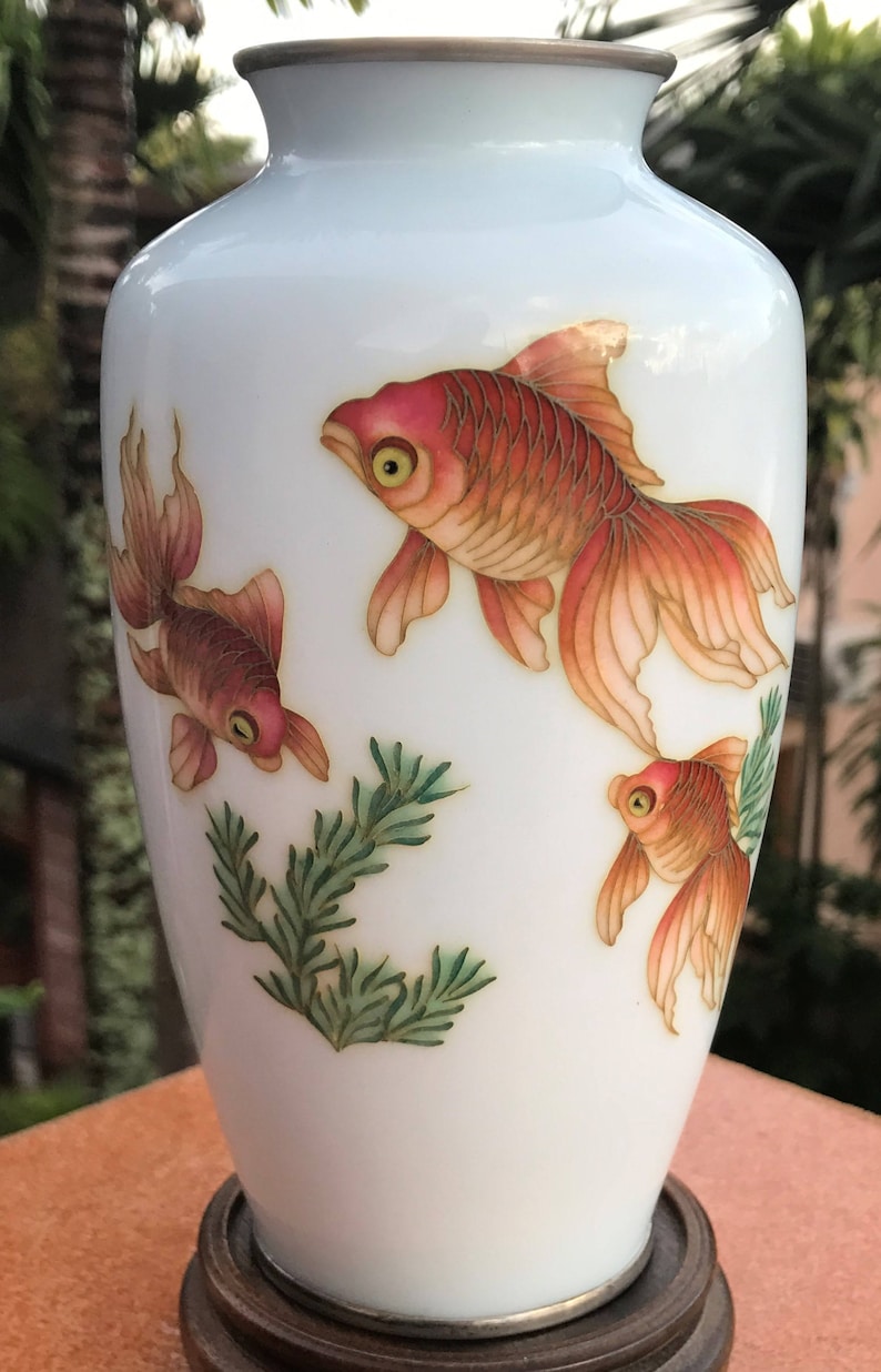Pu&ograve; includere: Vaso in porcellana bianca con bordo argentato e tre pesci dorati dipinti sul davanti. I pesci nuotano in un disegno floreale verde e bianco.