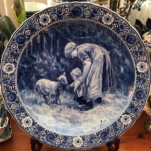 May include: A blue and white Delftware plate featuring a scene of two figures and a lamb in a woodland setting. The plate has a decorative border with floral and scroll designs.