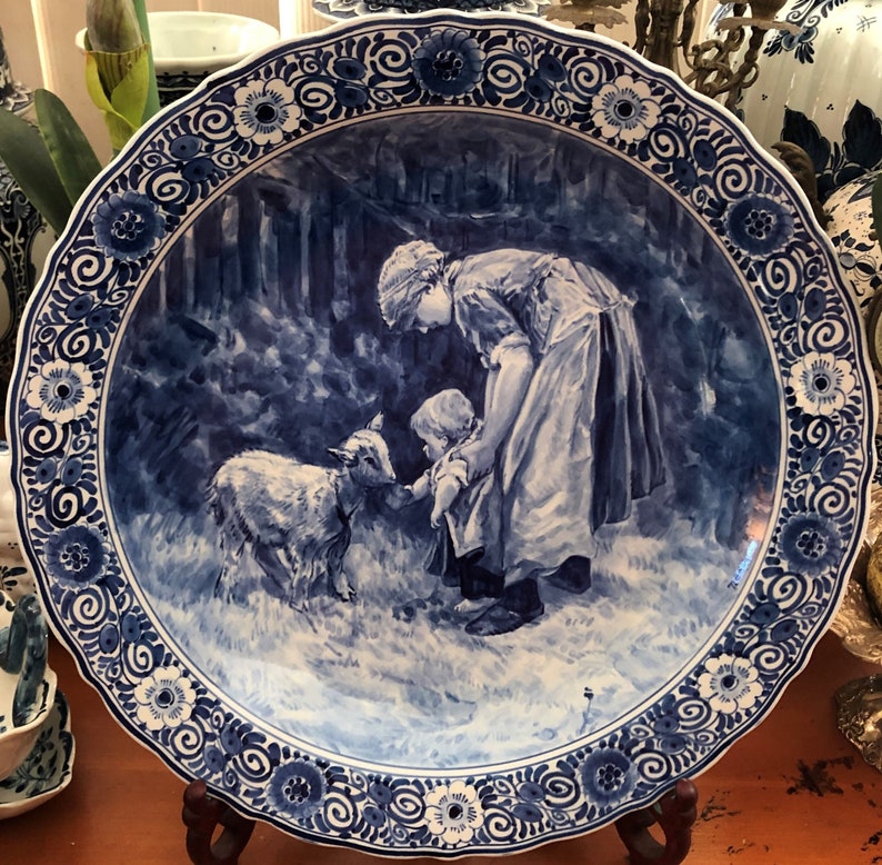 May include: A blue and white Delftware plate featuring a scene of two figures and a lamb in a woodland setting. The plate has a decorative border with floral and scroll designs.