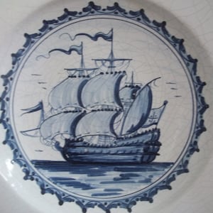 May include: White ceramic plate with a hand-painted design of a sailing ship in shades of blue. The ship is centered within a circular frame, surrounded by a decorative border. The plate has a slightly cracked appearance.