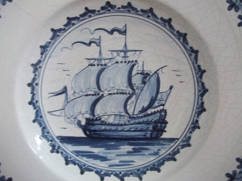 May include: White ceramic plate with a hand-painted design of a sailing ship in shades of blue. The ship is centered within a circular frame, surrounded by a decorative border. The plate has a slightly cracked appearance.