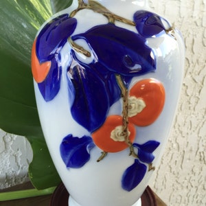 May include: A white porcelain vase with a gold rim, decorated with blue and orange stylized flowers and leaves. The vase is sitting on a wooden base.