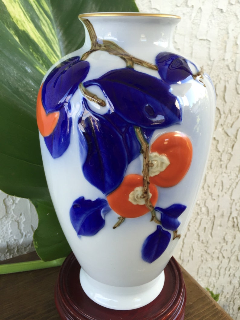 May include: A white porcelain vase with a gold rim, decorated with blue and orange stylized flowers and leaves. The vase is sitting on a wooden base.