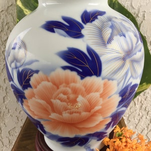 May include: A white ceramic vase with a gold rim, featuring a large peach-coloured peony and blue floral accents. The vase has a round shape and sits on a wooden base, showcasing a classic floral design.