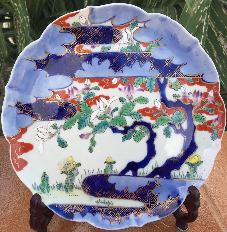 May include: A decorative ceramic plate with a scalloped edge. The plate features a landscape design with a blue tree, white flowers, and red and blue accents. Gold detailing adds to the ornate design. The plate is displayed on a wooden stand.