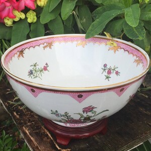 Op de afbeelding: Een grote witte porseleinen kom met een roze en gouden rand en een bloemenpatroon. De kom staat op een houten standaard.