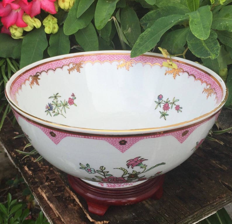Op de afbeelding: Een grote witte porseleinen kom met een roze en gouden rand en een bloemenpatroon. De kom staat op een houten standaard.