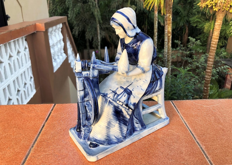 K&ouml;nnte beinhalten: Blau-wei&szlig;e Keramikfigur, die eine Frau an einem Spinnrad darstellt. Die Figur hat ein traditionelles niederl&auml;ndisches Design, mit einer Frau, die eine Haube und eine Sch&uuml;rze tr&auml;gt. Die Frau sitzt auf einem Stuhl.