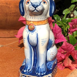 K&ouml;nnte beinhalten: Keramikhundfigur mit wei&szlig;em Grund und blauen Akzenten. Der Hund hat blaue Ohren, ein blaues Halsband mit orangefarbenem Mittelteil und ein l&auml;chelndes Gesicht. Die Figur steht auf einem dekorativen Sockel mit floralen Mustern.