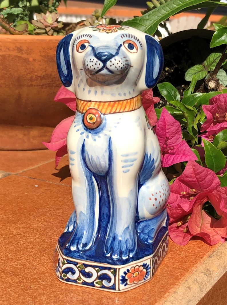 K&ouml;nnte beinhalten: Keramikhundfigur mit wei&szlig;em Grund und blauen Akzenten. Der Hund hat blaue Ohren, ein blaues Halsband mit orangefarbenem Mittelteil und ein l&auml;chelndes Gesicht. Die Figur steht auf einem dekorativen Sockel mit floralen Mustern.