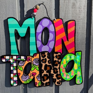 May include: A colorful wooden sign spelling "Montana" with a variety of patterns and colors. The letters are painted in teal stripes, purple polka dots, orange and pink stripes, floral designs, leopard print, and green with small colorful shapes.