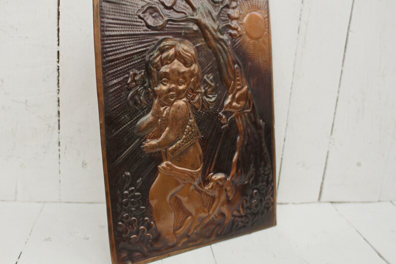 May include: A copper wall plaque with a raised relief design of a child holding a flower and standing next to a tree. The plaque has a sun and a floral design in the background.