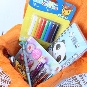 K&ouml;nnte beinhalten: Ein Geschenkkorb gef&uuml;llt mit bunten Bastelutensilien, darunter Werkzeuge zum Modellieren von Ton, ein Notizbuch mit einem Pandab&auml;r, Glitzerkleber und Radiergummis. Der Korb ist mit orangefarbenem Seidenpapier ausgelegt.