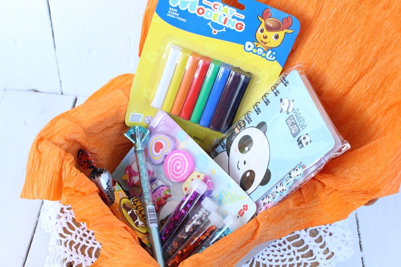 K&ouml;nnte beinhalten: Ein Geschenkkorb gef&uuml;llt mit bunten Bastelutensilien, darunter Werkzeuge zum Modellieren von Ton, ein Notizbuch mit einem Pandab&auml;r, Glitzerkleber und Radiergummis. Der Korb ist mit orangefarbenem Seidenpapier ausgelegt.