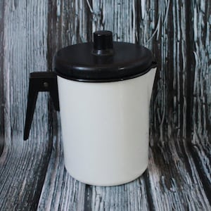 May include: A white enamel coffee pot with a black lid and handle. The pot is cylindrical with a pouring spout. The handle is attached to the side. The lid has a black knob. A vintage kitchenware item.