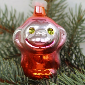 Puede incluir: Un adorno de Navidad de vidrio rojo y plateado con forma de mono con ojos verdes. El adorno está colgado de una rama de pino verde.