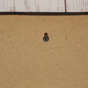 May include: A close-up of a brown cardboard backing with a black metal hook in the center. The backing is framed with a black painted wooden frame.