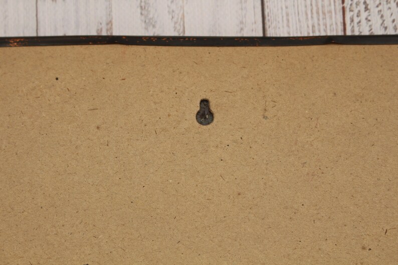 May include: A close-up of a brown cardboard backing with a black metal hook in the center. The backing is framed with a black painted wooden frame.