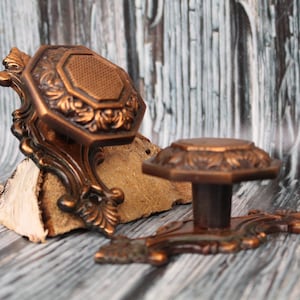 Può includere: Due pomelli decorativi per armadi, di colore rame, con intricati disegni floreali. I pomelli sono su una superficie di legno.