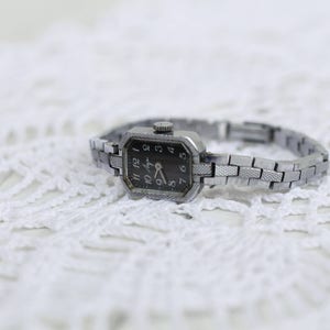 K&ouml;nnte beinhalten: Eine silberne Armbanduhr mit schwarzem Zifferblatt und silbernem Metallband. Die Uhr hat ein rechteckiges Zifferblatt mit r&ouml;mischen Ziffern.