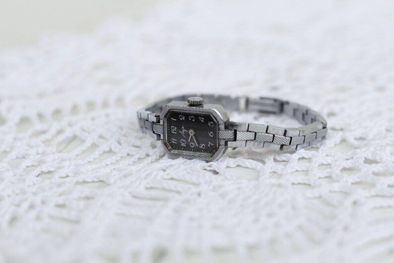K&ouml;nnte beinhalten: Eine silberne Armbanduhr mit schwarzem Zifferblatt und silbernem Metallband. Die Uhr hat ein rechteckiges Zifferblatt mit r&ouml;mischen Ziffern.