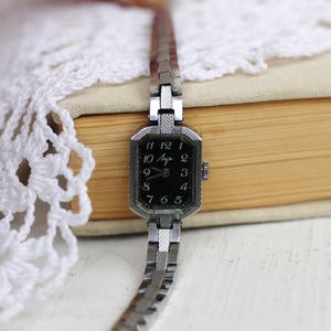 K&ouml;nnte beinhalten: Eine silberne Armbanduhr mit schwarzem Zifferblatt und wei&szlig;en Zahlen. Die Uhr hat ein silbernes Armband und ein quadratisches Geh&auml;use. Auf dem Zifferblatt der Uhr steht "Anya".