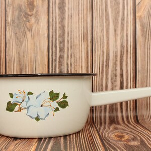 Puede incluir: Cacerola de esmalte blanco con borde negro y asa blanca. La cacerola tiene un diseño floral con flores azules y hojas verdes.