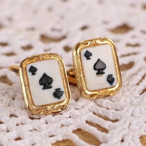 May include: A pair of gold cufflinks with a white background and a black spade design. The cufflinks are on a white crocheted surface.