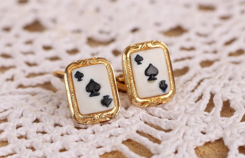 May include: A pair of gold cufflinks with a white background and a black spade design. The cufflinks are on a white crocheted surface.