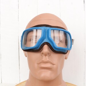 May include: Blue safety goggles with a black strap and clear lenses. The goggles are on a mannequin head.