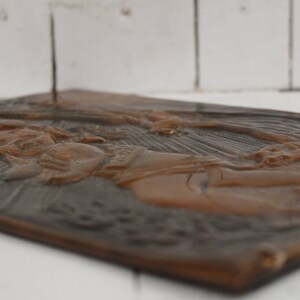 May include: A brown metal plaque with a raised relief design of a woman holding a child. The plaque has a textured surface and a dark brown patina.