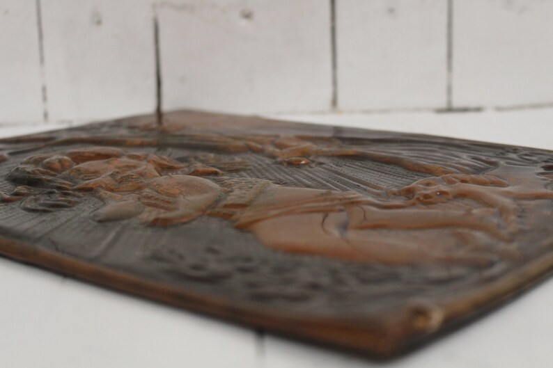 May include: A brown metal plaque with a raised relief design of a woman holding a child. The plaque has a textured surface and a dark brown patina.