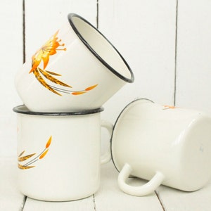 May include: Three white enamel mugs with black rims and a floral design in shades of orange and yellow. The mugs are stacked on top of each other.