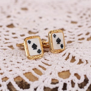May include: A pair of gold cufflinks with a white background and black spades. The cufflinks are on a white crocheted surface.