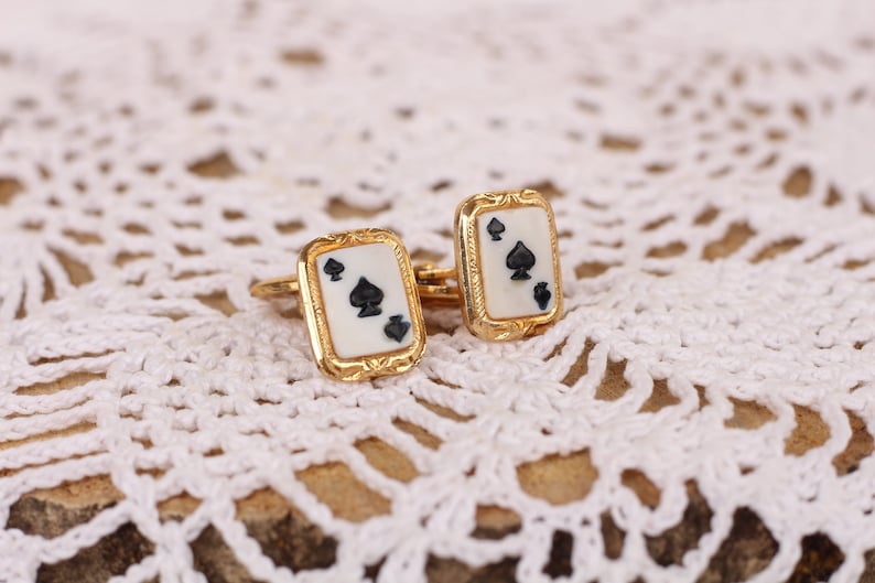 May include: A pair of gold cufflinks with a white background and black spades. The cufflinks are on a white crocheted surface.