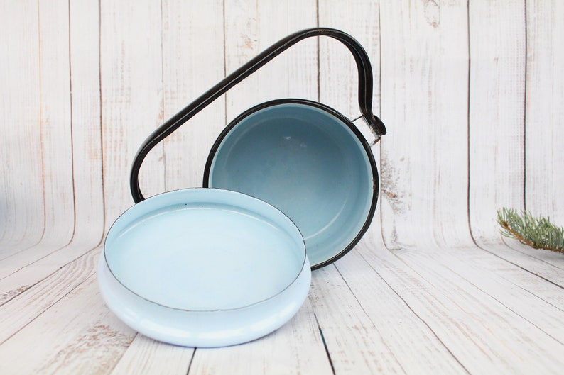 Op de afbeelding: Een lichtblauwe emaille pan met een zwarte handgreep en deksel. De pan is open en het deksel ligt ernaast.