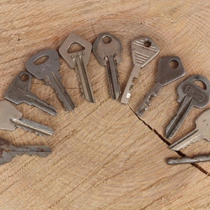 May include: A collection of nine vintage keys arranged in a semi-circle on a wooden surface. The keys are made of metal and have different shapes and sizes. Some of the keys have visible markings and numbers.