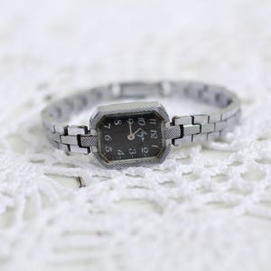 K&ouml;nnte beinhalten: Eine silberne Armbanduhr mit schwarzem Zifferblatt und silbernem Metallband. Die Uhr hat ein rechteckiges Zifferblatt mit leicht abgerundeter Kante. Das Zifferblatt der Uhr hat schwarze Zahlen und Zeiger.