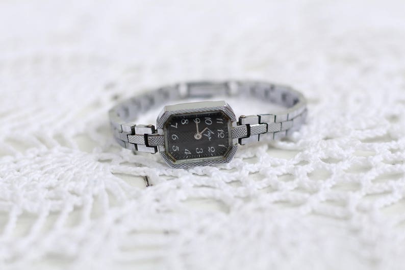 K&ouml;nnte beinhalten: Eine silberne Armbanduhr mit schwarzem Zifferblatt und silbernem Metallband. Die Uhr hat ein rechteckiges Zifferblatt mit leicht abgerundeter Kante. Das Zifferblatt der Uhr hat schwarze Zahlen und Zeiger.