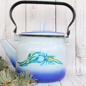 Op de afbeelding: Een blauwe en witte emaille theepot met een zwart handvat en een bloemenontwerp. De theepot staat op een wit houten oppervlak.